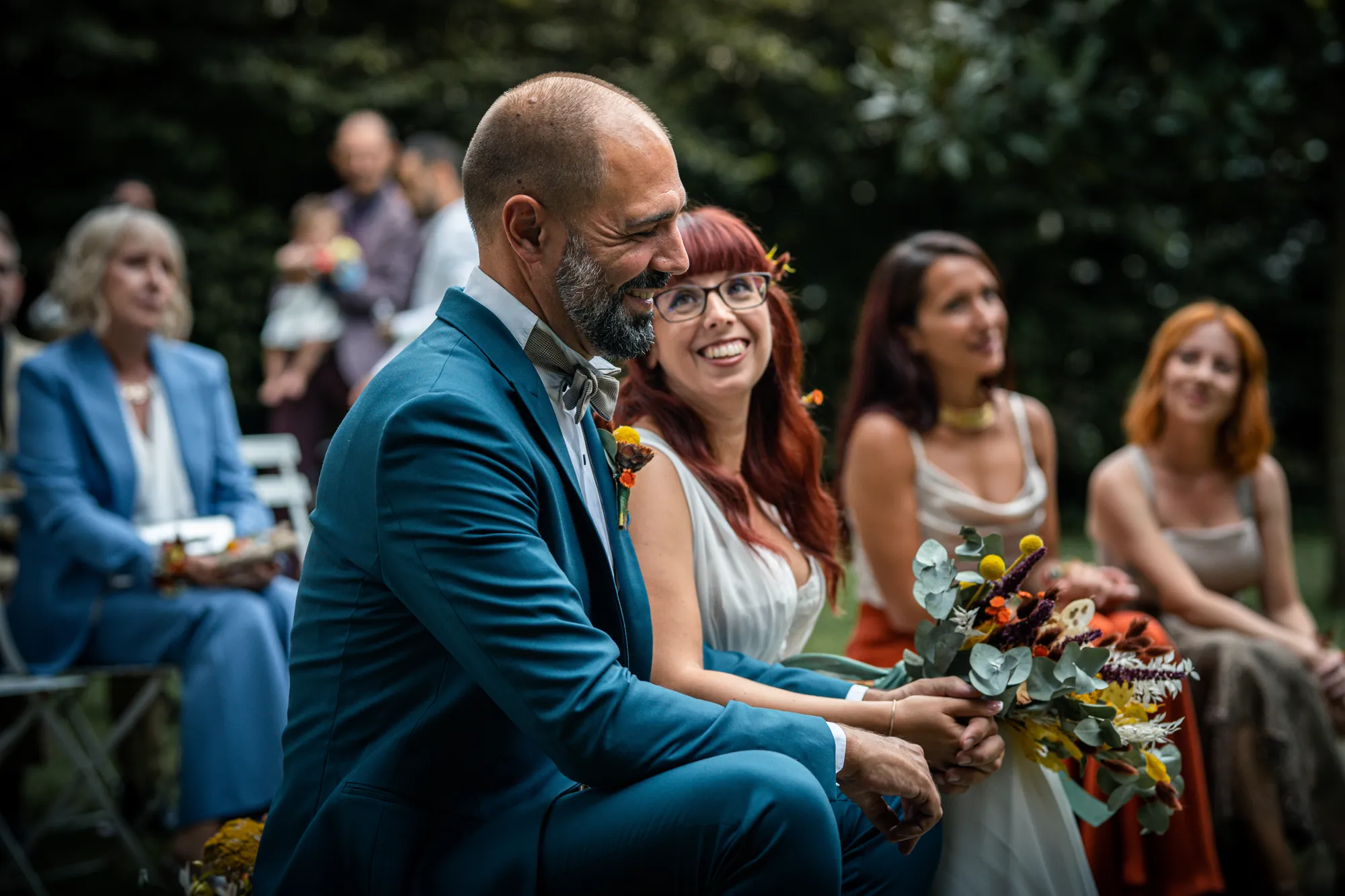 Sposi seduti durante la cerimonia, sorrisi e bouquet colorato